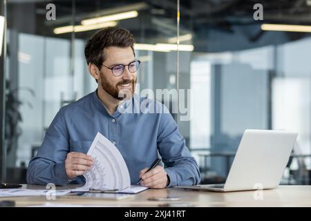 Ein lächelnder junger Mann sitzt im Büro an einem Tisch vor einem Laptop, macht Notizen in Dokumenten, arbeitet mit Daten. Stockfoto