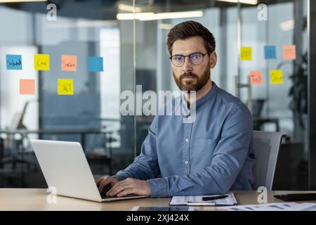 Porträt eines jungen selbstbewussten und ernstzunehmenden jungen Geschäftsmannes, der in einem modernen Büro am Schreibtisch sitzt, mit einem Laptop arbeitet und auf die Kamera blickt. Stockfoto