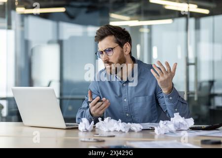 Verärgerter und enttäuschter junger Mann, der im Büro sitzt und mit einem Videoanruf auf dem Laptop spricht, zerknittertes Papier auf dem Tisch, gestikuliert mit den Händen. Stockfoto