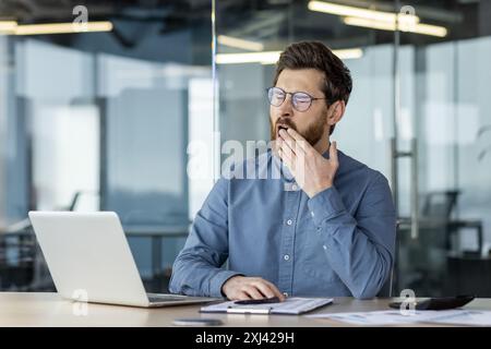 Ein müder junger Geschäftsmann sitzt im Büro, arbeitet Überstunden am Laptop, gähnt, bedeckt seinen Mund mit der Hand. Stockfoto
