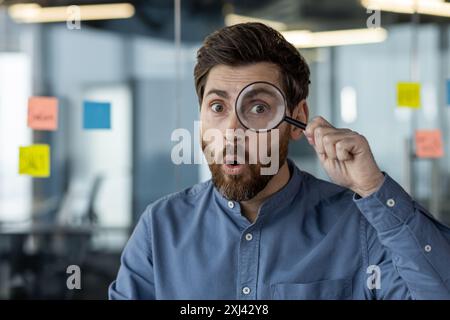 Nahaufnahme eines jungen Mannes im Büro, der durch eine Lupe überrascht auf die Kamera blickt. Stockfoto