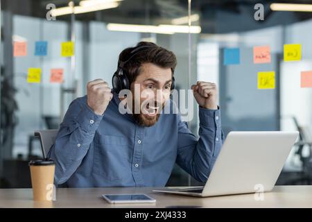 Ein glücklicher junger Mann mit Headset sitzt im Büro am Schreibtisch und freut sich über Sieg und Erfolg, schaut auf den Bildschirm und hob die Hände hoch. Stockfoto