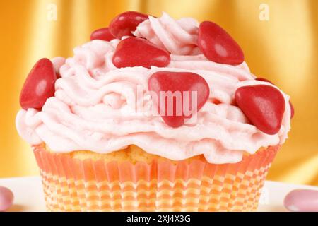 Fantasievoller Valentinstag-Cupcake, dekoriert mit kleinen roten, zuckerüberzogenen Schokoladenherzen auf einer weißen Platte, auf gelbem Hintergrund. Selektiver Fokus Stockfoto