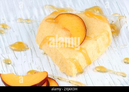 Herzförmiges bayerisches Pfirsich-Creme-Dessert mit Pfirsichsauce und Pfirsichscheiben auf einem transparenten Glasteller. Selektiver Fokus Stockfoto