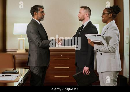 Seitenansicht zweier erfolgreicher Geschäftsleute in formalwear, die sich nach Vertragsabschluss anschauen und die Hand schütteln Stockfoto