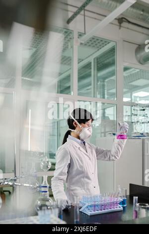 Seitenansicht einer jungen brünetten Forscherin im Schutzatemgerät beim Blick auf die violette Flüssigkeit im Reagenzglas während des Stehens im Labor Stockfoto