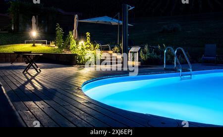 Luxuriöse Umgebung im Freien bei Nacht mit einem brillant beleuchteten Pool mit LED-Leuchten, die ein ruhiges blaues Leuchten erzeugen, umgeben von einem beleuchteten Garten Stockfoto