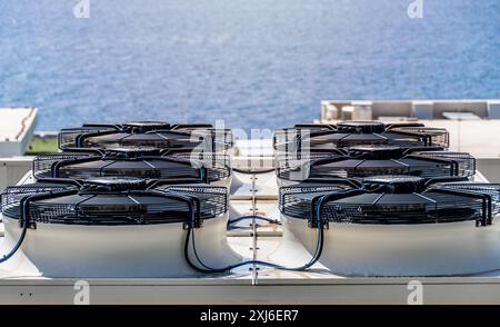 Sechs große schwarze Klimaanlagen, die auf einem Dach mit malerischem Meeresblick installiert sind. Effiziente Industriekühlungstechnologie spiegelt sich in wider Stockfoto