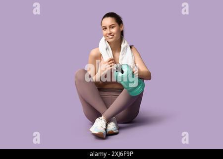 Junge sportliche Frau, die mit Wasserflasche und Handtuch auf Fliederhintergrund sitzt Stockfoto