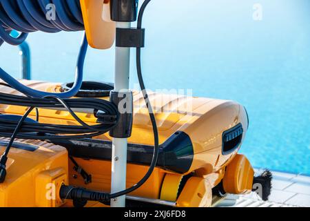 Nahaufnahme eines High-Tech-Roboter-Schwimmbadreinigers, ausgestattet mit Bürsten und einem langen Kabel, am Rand eines hellblauen Schwimmbades, reflekti Stockfoto