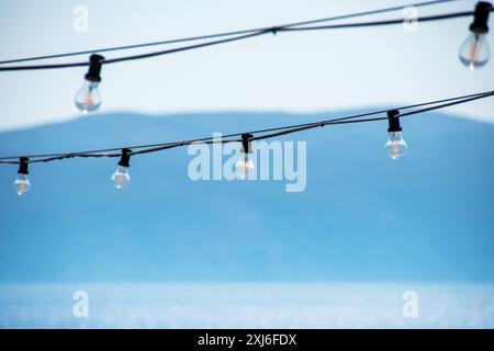 Klare Edison-Glühbirnen, die an Kabeln aufgehängt sind, stehen im Kontrast zu einem sanften blauen Himmel und einer fernen Bergkulisse. Das Bild fängt perfekt die Eleganz von uns ein Stockfoto