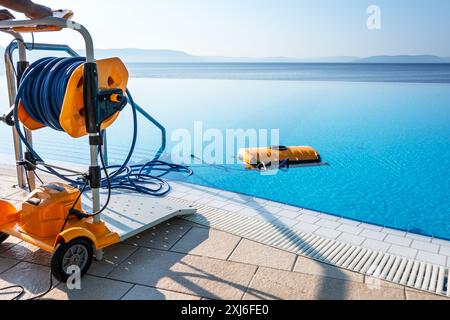 Sommerszene im Freien, mit einem Roboter-Poolreiniger in einem leuchtend blauen Infinity-Pool, ergänzt durch einen Schlauchwagen und ruhigen Meerblick, Highlight Stockfoto