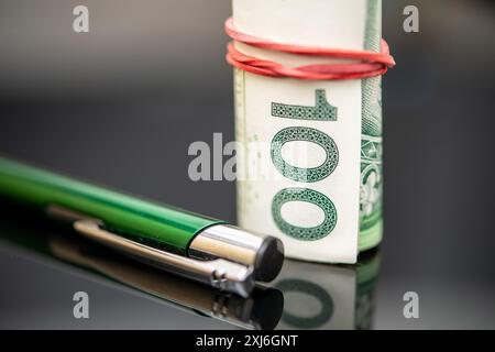 Detaillierte Ansicht von 100 polnischen Zlotys-Banknoten, die von einem roten Gummiband gerollt und gebunden sind, gepaart mit einem eleganten grünen Stift, der auf einer reflektierenden dunklen Oberfläche liegt; Stockfoto