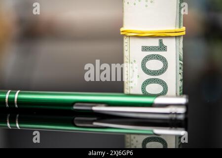 Nahaufnahme mit gerollten 100 polnischen Zloty-Banknoten, die durch ein gelbes Gummiband gesichert sind und neben zwei grünen Stiften auf einer reflektierenden Oberfläche liegen. Idee Stockfoto