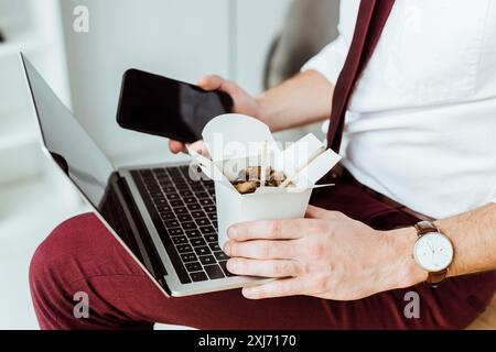 7/8-Ansicht des Geschäftsmannes Holding Box mit Nudeln, Smartphone und Laptop Stockfoto