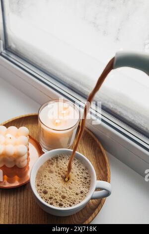 Eine gemütliche Szene mit Kaffee, der in eine Tasse auf einem Holztablett gegossen wird, neben zündeten Kerzen an einem frostigen Fenster. Stockfoto