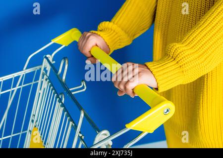 7/8-Ansicht der Frau in gelber Kleidung holding Warenkorb auf blauem Hintergrund Stockfoto