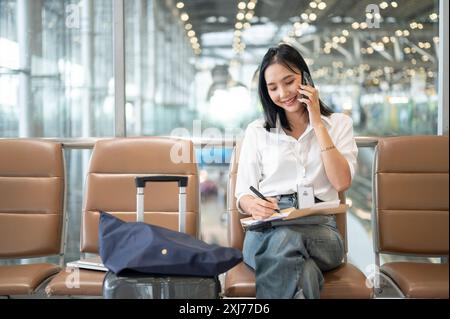 Eine selbstbewusste, beschäftigte asiatische Geschäftsfrau spricht am Telefon und macht Notizen in einer Zeitung, während sie im Flughafenterminal auf ihre Einstiegszeit wartet. Stockfoto