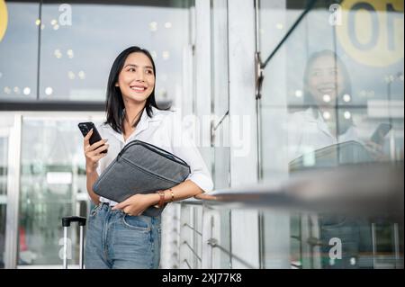 Eine attraktive asiatische Passagierin mit einem Smartphone in der Hand steht mit ihren Sachen an der Ausgangstür des Flughafenterminals und wartet auf sie Stockfoto