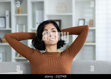 Nahaufnahme eines Porträts einer entspannten jungen Frau, die zu Hause auf der Couch sitzt und sich ausruht, Hände hinter ihrem Kopf, die Kamera ansieht und lächelt. Stockfoto