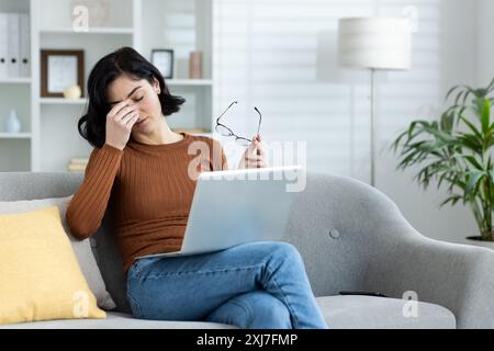 Müde junge Studentin, Freiberuflerin sitzt zu Hause auf der Couch, hält Brille und Laptop auf dem Schoß, wischt die Augen mit der Hand, arbeitet und studiert aus der Ferne und Überstunden. Stockfoto