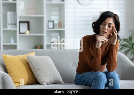 Eine junge Frau mit Schmerzen sitzt zu Hause auf dem Sofa und hält den Kopf mit der Hand, spürt Druck und Krämpfe, gibt eine Massage. Stockfoto