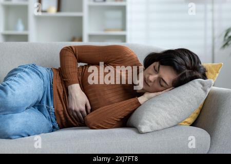 Frau mit braunem Pullover und Jeans, die auf einer Couch liegt und Bauchschmerzen in einer gemütlichen Wohnzimmerumgebung hat. Stockfoto