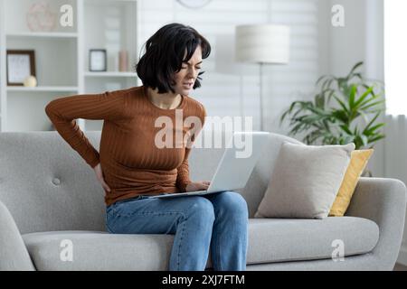 Junge Frau, die an Rückenschmerzen leidet, sitzt zu Hause auf dem Sofa, arbeitet und studiert ferngesteuert per Laptop und hält ihre Hand auf dem unteren Rücken. Stockfoto