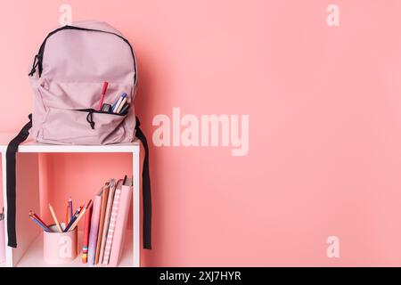 Es gibt Schulbedarf und einen Rucksack auf weißen Regalen vor rosafarbenem Hintergrund Stockfoto