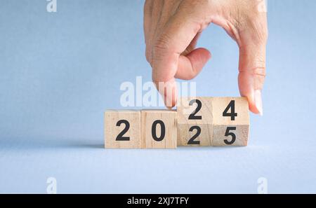 Finger drehen sich von 2024 auf 2025, Zahlen auf Holzblöcken. Auf blauem Hintergrund, Neujahrskonzept. Stockfoto