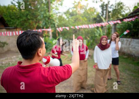 Ein Mann in einem roten Hemd hält ein Megaphon und zeigt auf eine Gruppe von Leuten, die ein Spiel spielen. Indonesisches Unabhängigkeitstag-Konzept Stockfoto