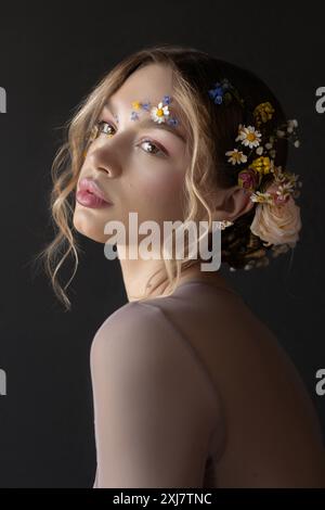 Eine junge Frau mit langen, blonden, lockigen Haaren, mit Blumen im Haar und im Gesicht, posiert für ein Atelierporträt. Stockfoto