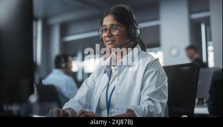 Porträt eines Smart Young Indian Specialist, der in einer technischen Startup-Helpdesk-Abteilung arbeitet und Computer in einem vielfältigen Büro verwendet. Frau trägt Brille und Kopfhörer mit Mikrofon Stockfoto