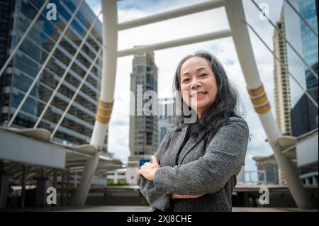 Eine selbstbewusste, reife asiatische Geschäftsfrau oder Geschäftsführerin in einem formellen Business-Anzug steht mit überkreuzten Armen in der großen Stadt und lächelt das Kommen an Stockfoto