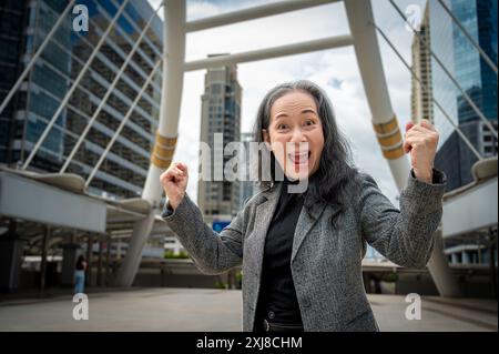 Eine aufgeregte, glückliche, reife asiatische Geschäftsfrau zeigt ihre triumphalen Fäuste, feiert ihre erfolgreiche Erreichung ihrer Ziele und steht in der Stadt Stockfoto