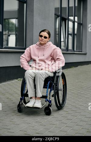 Eine junge Frau sitzt in einem Rollstuhl auf dem Bürgersteig vor einem Gebäude. Sie trägt einen rosafarbenen Hoodie und eine Sonnenbrille. Stockfoto