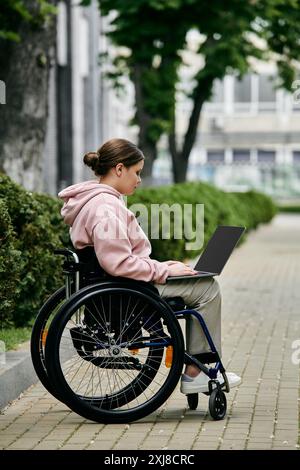 Eine junge Frau mit Behinderung sitzt in einem Rollstuhl auf einem gepflasterten Gehweg, mit einem Laptop. Stockfoto