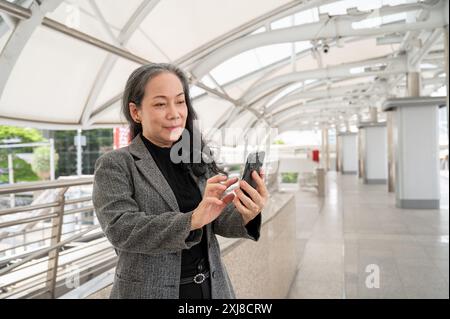 Eine glückliche, attraktive, reife asiatische Geschäftsfrau in einem Anzug liest SMS oder liest Nachrichten auf ihrem Smartphone, während sie auf einem Skywalk oder einem Skyt steht Stockfoto