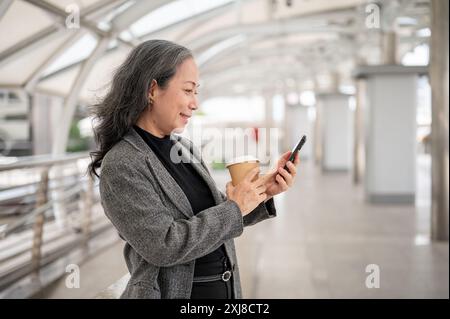 Eine glückliche, attraktive, reife asiatische Geschäftsfrau in einem Anzug liest SMS oder liest Nachrichten auf ihrem Smartphone, während sie auf einem Skywalk oder einem Skyt steht Stockfoto