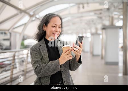 Eine glückliche, attraktive, reife asiatische Geschäftsfrau in einem Anzug liest SMS oder liest Nachrichten auf ihrem Smartphone, während sie auf einem Skywalk oder einem Skyt steht Stockfoto