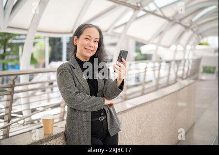Eine attraktive, reife asiatische Geschäftsfrau im Business-Anzug steht mit einem Smartphone in der Hand auf einem Skywalk und lächelt in die Kamera. Geschäftsleute A Stockfoto