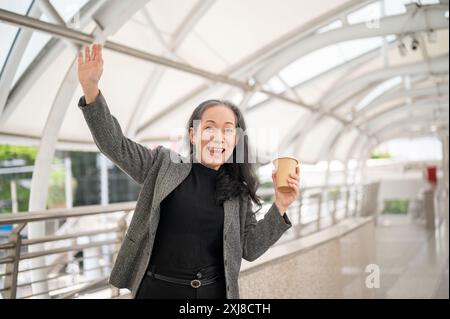 Eine fröhliche, reife asiatische Geschäftsfrau in einem Business-Anzug winkt mit der Hand und lächelt in die Kamera und grüßt jemanden, während sie auf einem Skywalk steht Stockfoto