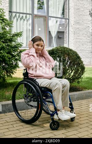 Die junge Frau im rosa Hoodie entspannt sich im Rollstuhl und hört Musik auf dem gepflasterten Gehweg. Stockfoto