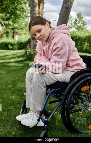 Eine junge Frau sitzt in einem Rollstuhl in einem grasbewachsenen Park und schaut auf ihr Handy herunter. Stockfoto