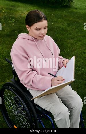 Eine junge Frau in einem rosafarbenen Hoodie sitzt in einem Rollstuhl auf einem grasbewachsenen Rasen, während sie in ein Notizbuch schreibt. Stockfoto