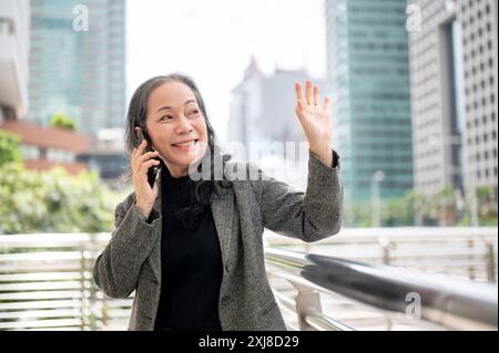 Eine glückliche, reife asiatische Geschäftsfrau spricht am Telefon und winkt mit der Hand, um jemanden zu begrüßen, während sie auf einem Skywalk oder im Freien steht, umgeben von Stockfoto