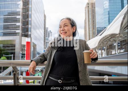 Eine selbstbewusste, positive, reife asiatische Geschäftsfrau steht auf einem Wolkenkratzer in der belebten Stadt. Stockfoto