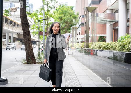 Eine selbstbewusste, positive, reife asiatische Geschäftsfrau läuft auf einem Bürgersteig in der Stadt, hält ihr Smartphone und eine Aktentasche und pendelt in die Arbeit Stockfoto