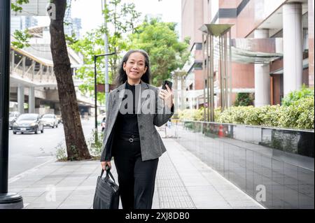 Eine selbstbewusste, positive, reife asiatische Geschäftsfrau läuft auf einem Bürgersteig in der Stadt, hält ihr Smartphone und eine Aktentasche und pendelt in die Arbeit Stockfoto