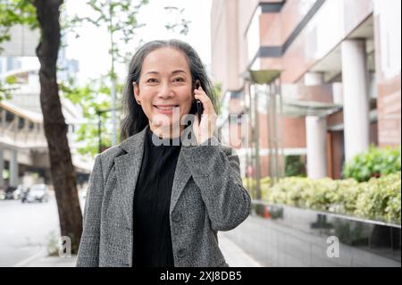 Eine schöne, positive, reife asiatische Geschäftsfrau telefoniert am Telefon, während sie ihre Arbeit pendelt und auf einem Bürgersteig in der Stadt läuft. Geschäftsleute Stockfoto